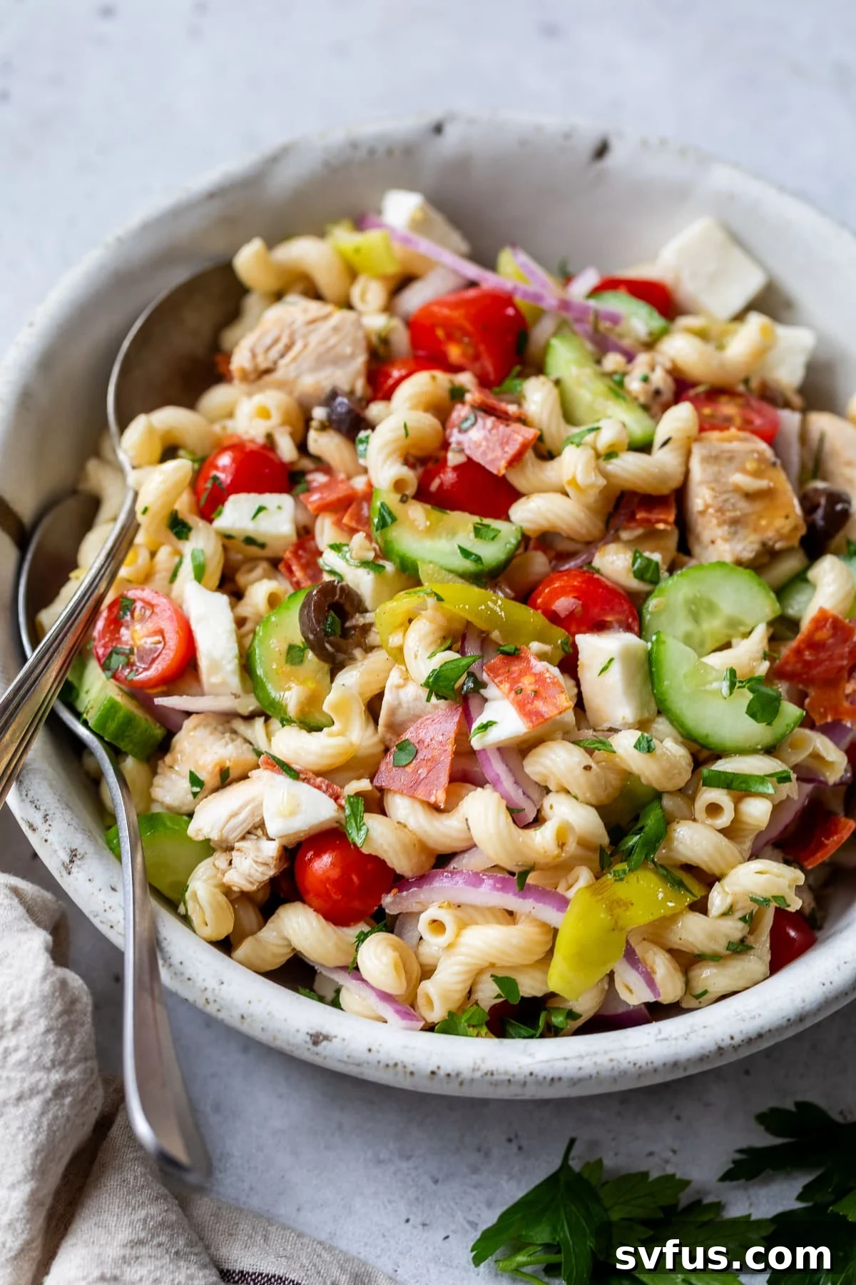 Büyük beyaz bir kasede iki kaşıkla servis edilen İtalyan makarna salatası.