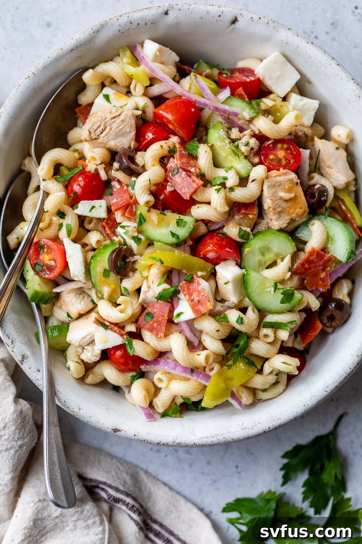 Büyük beyaz bir kasede iki kaşıkla servis edilen İtalyan makarna salatası.