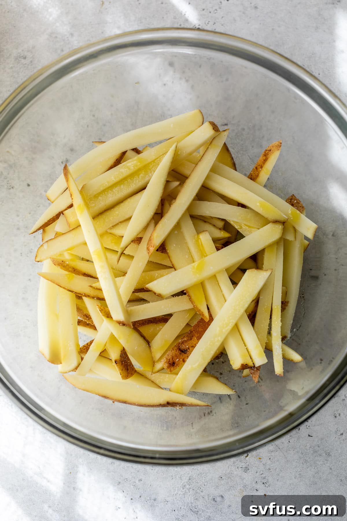Raw french fries seasoned with salt and olive oil in a clear glass bowl, ready for the air fryer.
