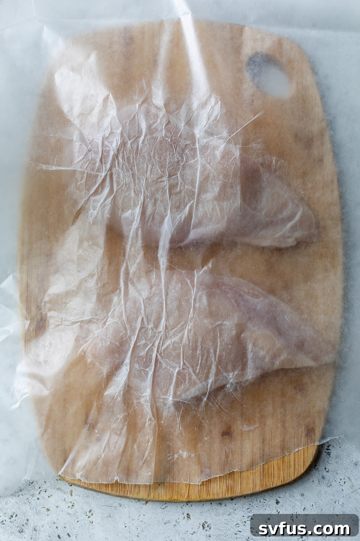Two boneless, skinless chicken breasts resting on a wooden cutting board, with a sheet of wax paper on top, ready for pounding to even thickness.
