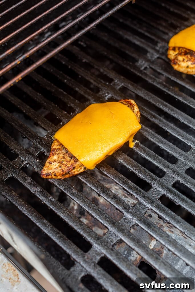 A perfectly grilled chicken breast, now topped with a slice of melted cheese, continuing to warm on an outdoor grill, ready for assembly.