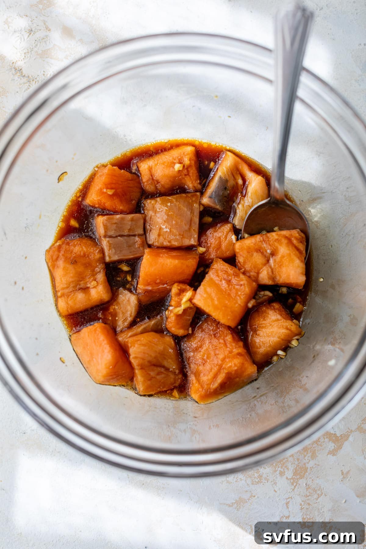 A spoon gently tosses vibrant salmon bites, glistening with a savory soy sauce marinade, in a clear glass mixing bowl, preparing them for the air fryer.