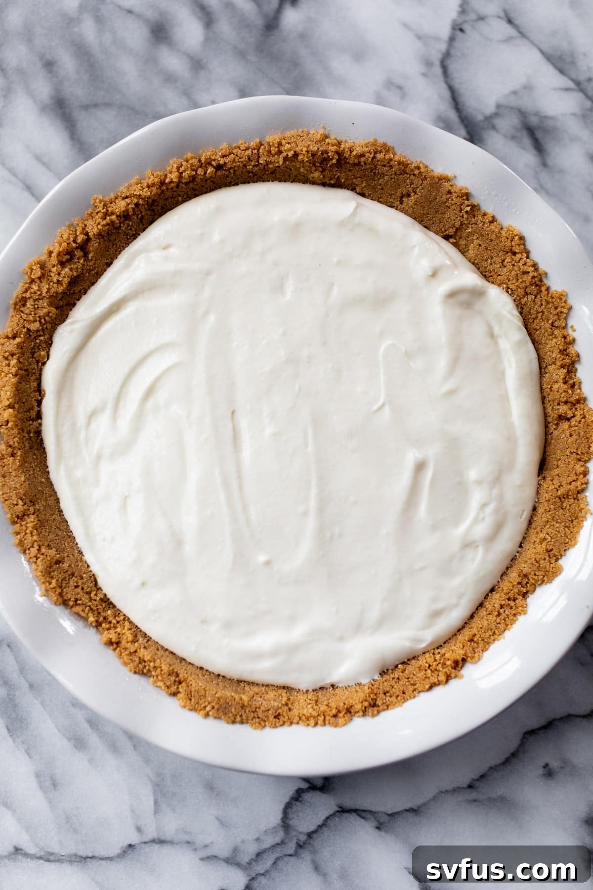 A chilled No Bake Cherry Cheesecake with a golden graham cracker crust and smooth white filling, waiting to be topped, sitting on a marble table.