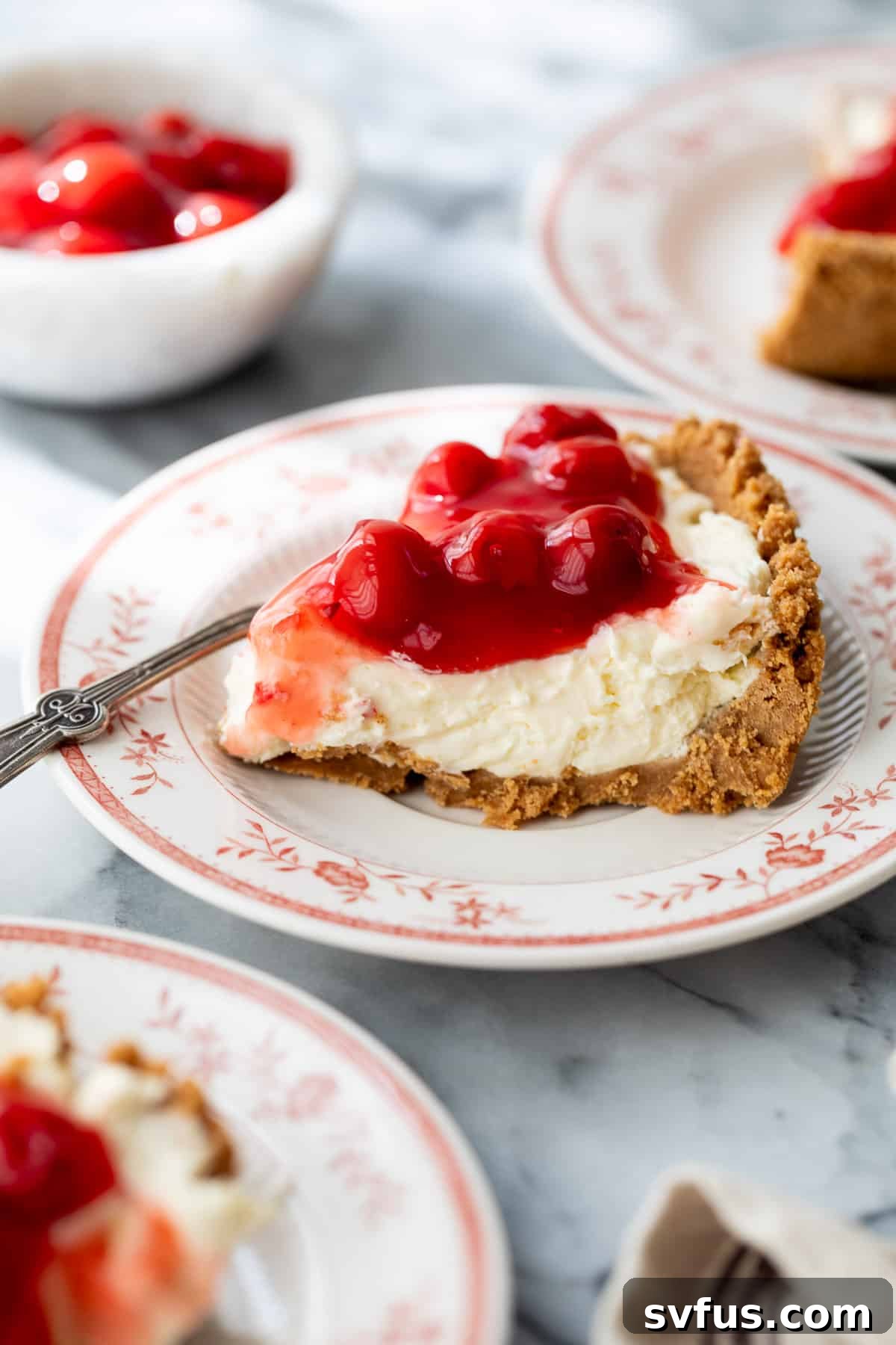 A perfect slice of No Bake Cherry Cheesecake on a white plate, topped with glistening whole cherries, ready to be enjoyed.