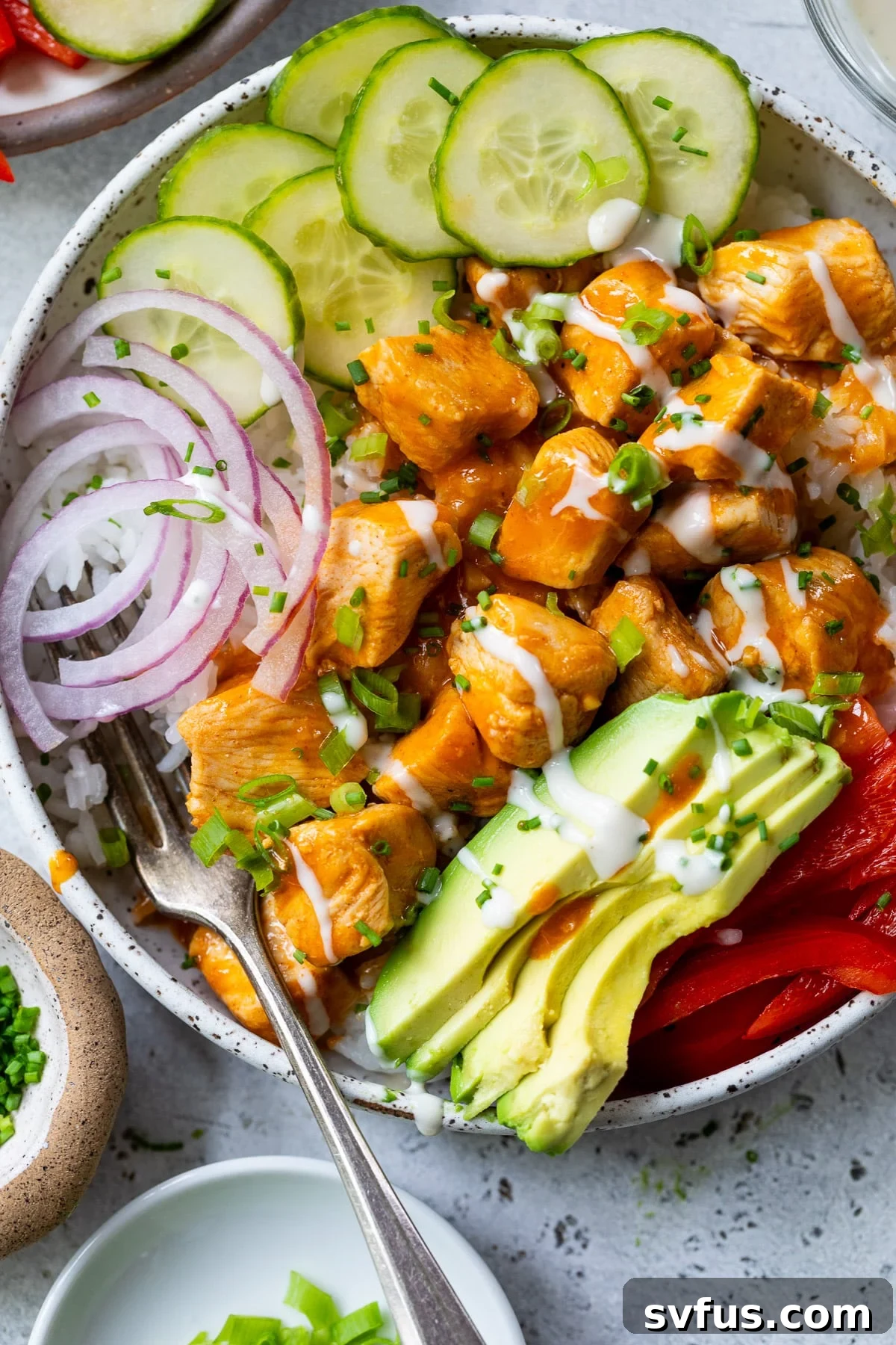 Overhead view of a vibrant Buffalo Chicken Bowl featuring succulent buffalo chicken bites, refreshing cucumber slices, thinly cut red onion, and creamy avocado chunks, all artfully arranged over a bed of rice.