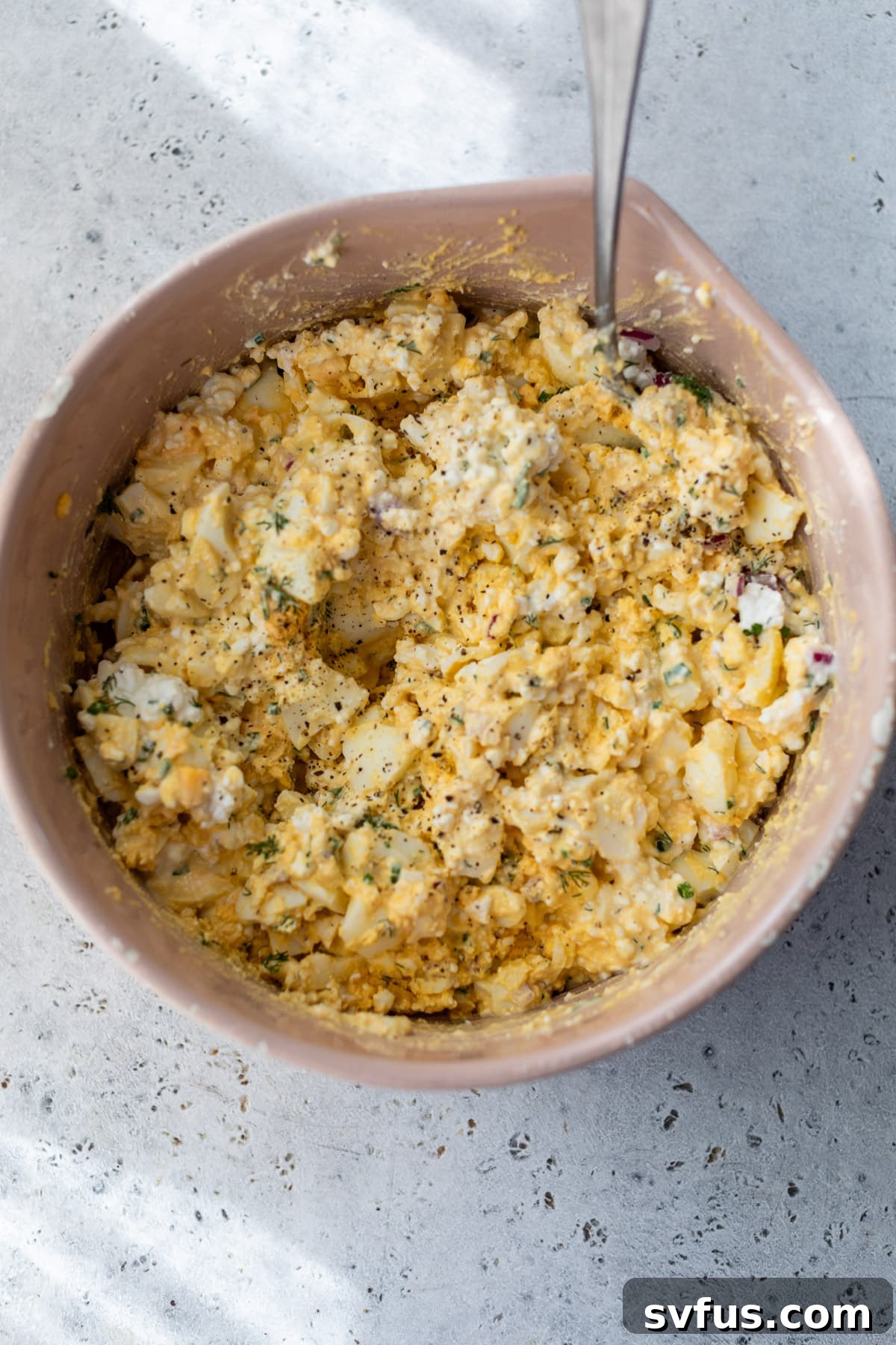 A spoon actively stirring creamy cottage cheese egg salad in a large pink bowl, showcasing the final mixed texture.