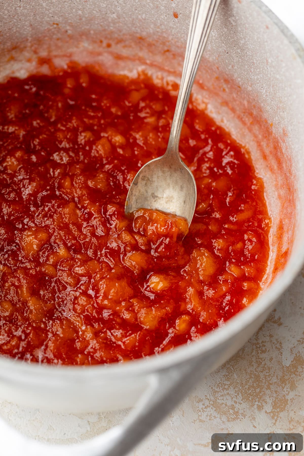 A wooden spoon stirring a pot of Peach Preserves, showing the chunky texture.