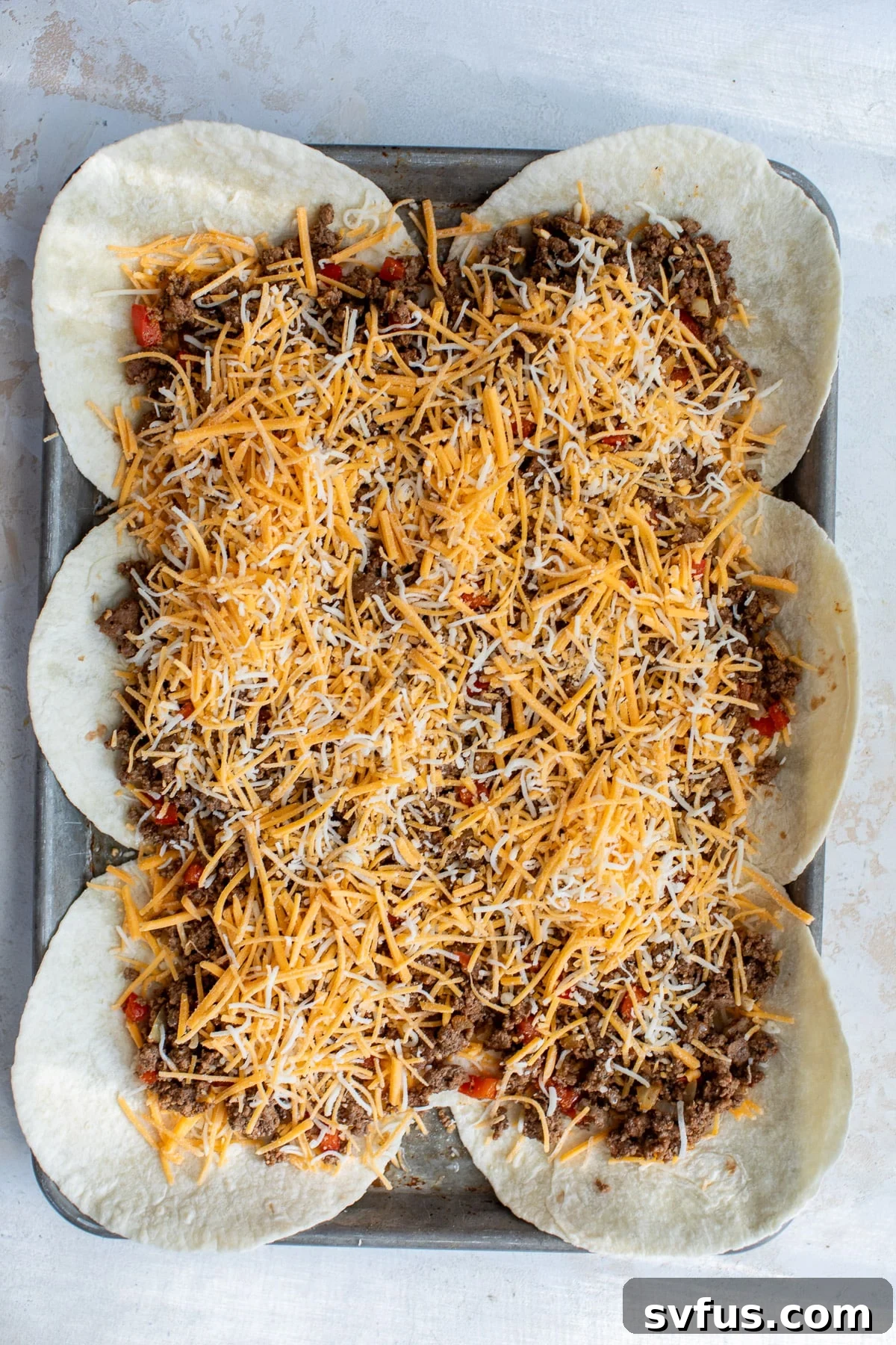 Layering the cooked ground beef filling and shredded cheese over the strategically placed tortillas on a sheet pan, ready for the next step.