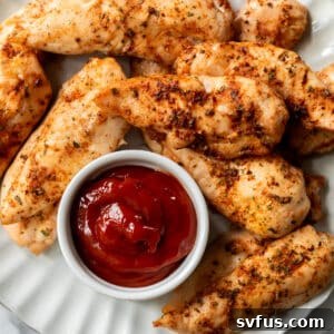 A pile of baked chicken tenders on a white plate next to a small bowl of ketchup.