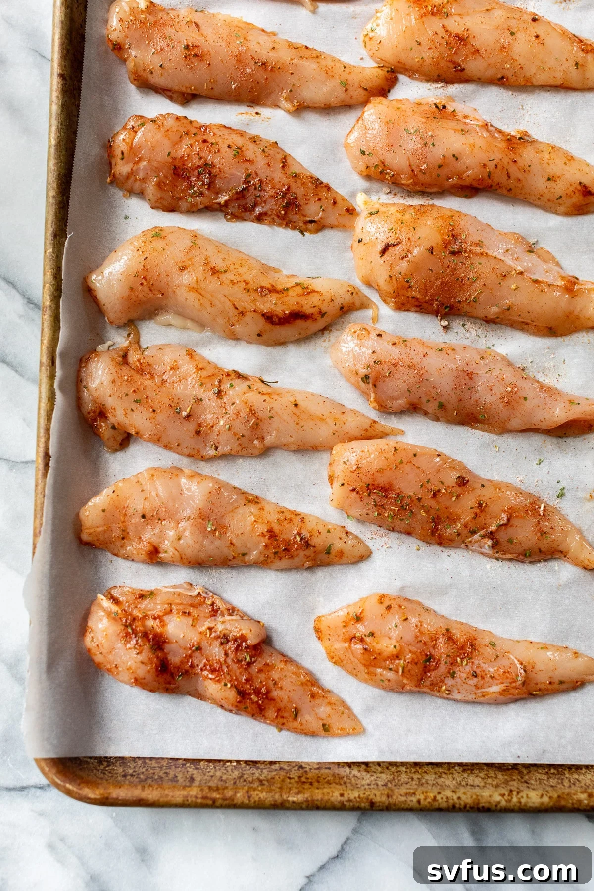 Raw, seasoned chicken tenders neatly arranged in rows on a parchment-lined baking sheet, ready for the oven.