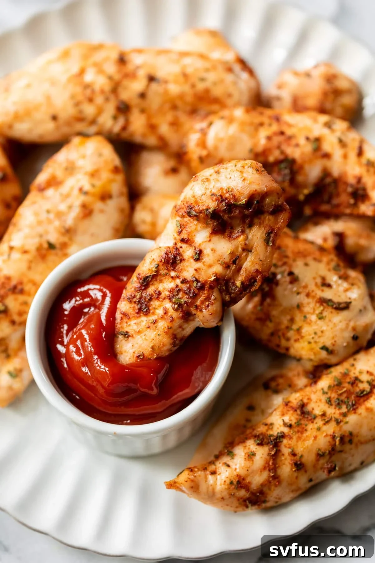 A hand gently dipping a golden-brown baked chicken tender into a small, inviting bowl of ketchup, showcasing the perfect pairing.