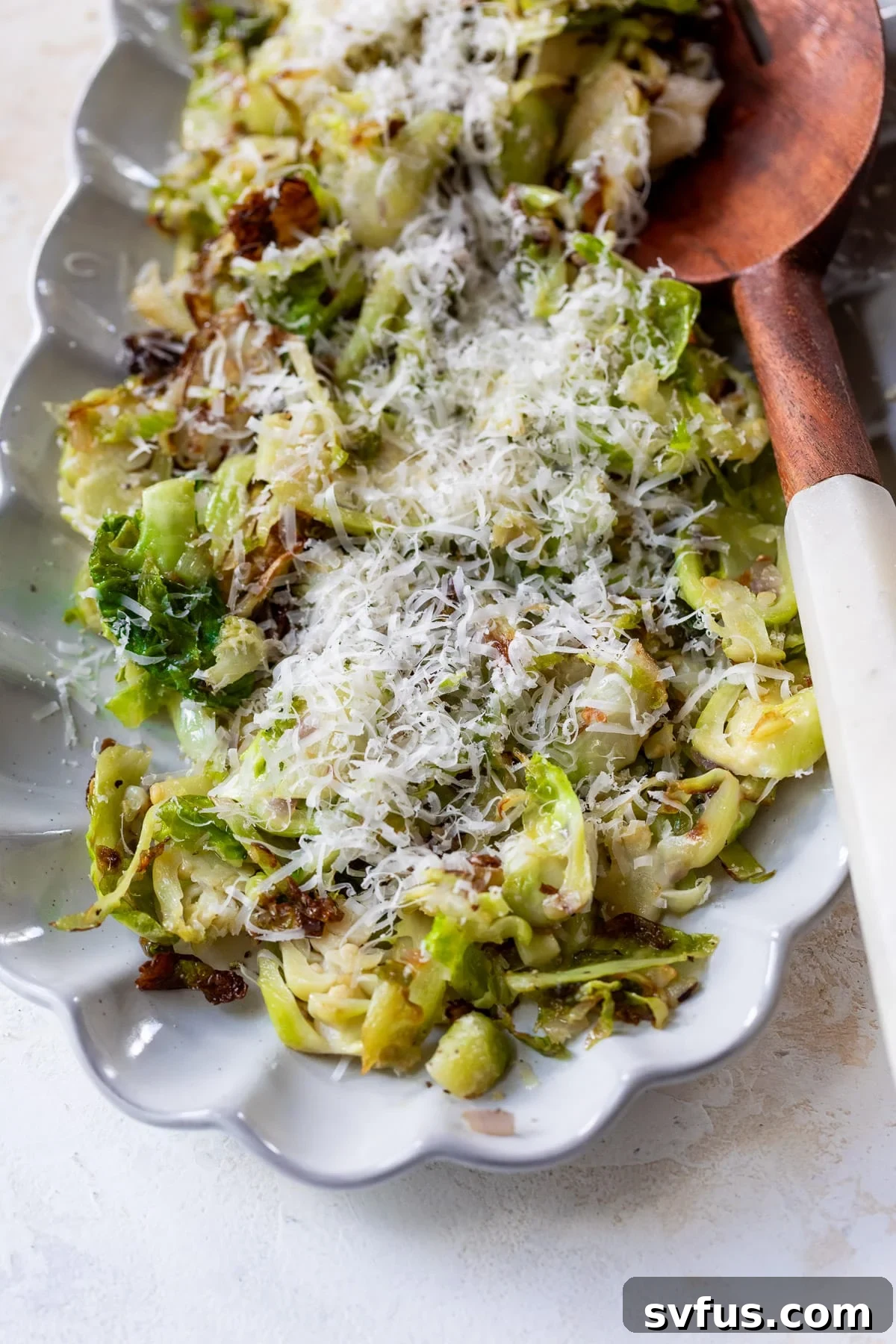 A serving platter filled with golden, crispy shaved Brussels sprouts, generously topped with shredded Parmesan cheese and garnished with a wooden spoon.