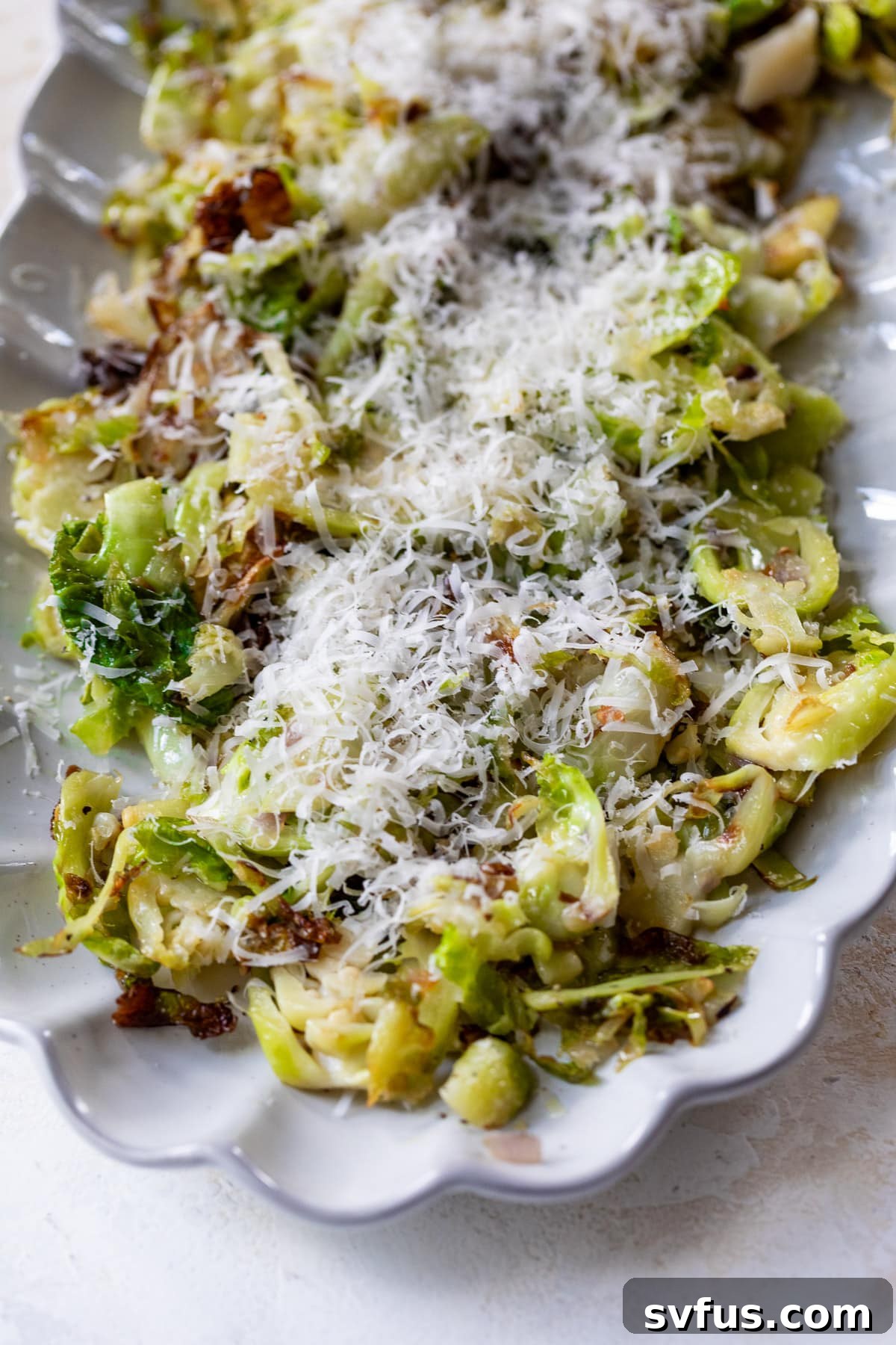 A white serving platter showcasing golden-brown shaved Brussels sprouts, topped with a generous layer of shredded Parmesan cheese, ready to be enjoyed.