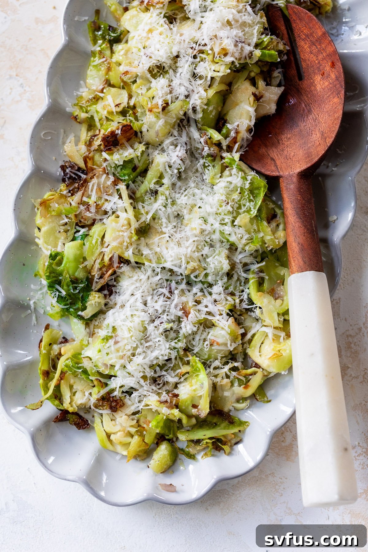 A final close-up of shaved Brussels sprouts garnished with fresh Parmesan cheese and a wooden spoon on a white serving platter, highlighting their crispy texture.