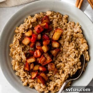 A gray colored bowl beside a cinnamon stick and a linen. Inside the bowl is cooked oatmeal topped with diced cooked apple.