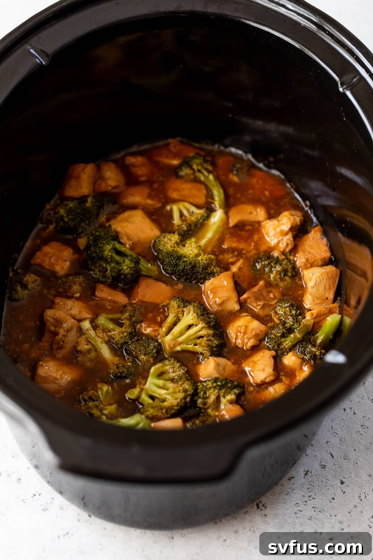 Close-up of chicken pieces and broccoli florets simmering in a rich orange sauce within a slow cooker.