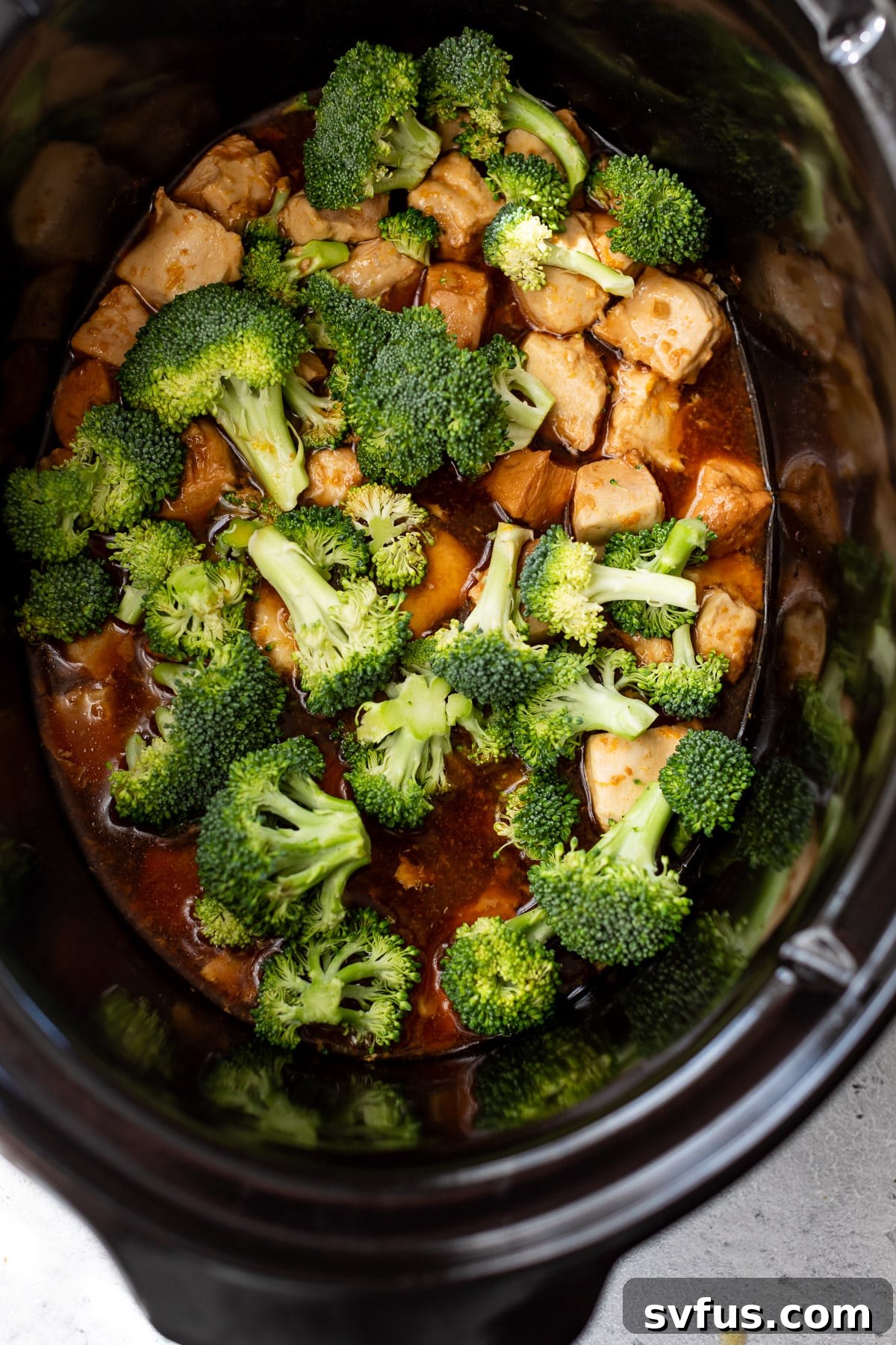 Broccoli florets and chicken pieces simmering in orange sauce in a slow cooker.