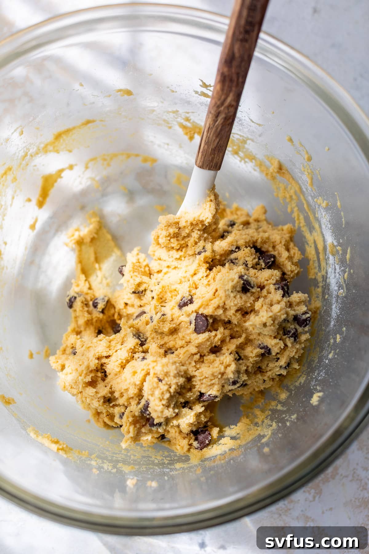 A spatula gently folding chocolate chips into the coconut flour cookie dough in a large glass bowl.