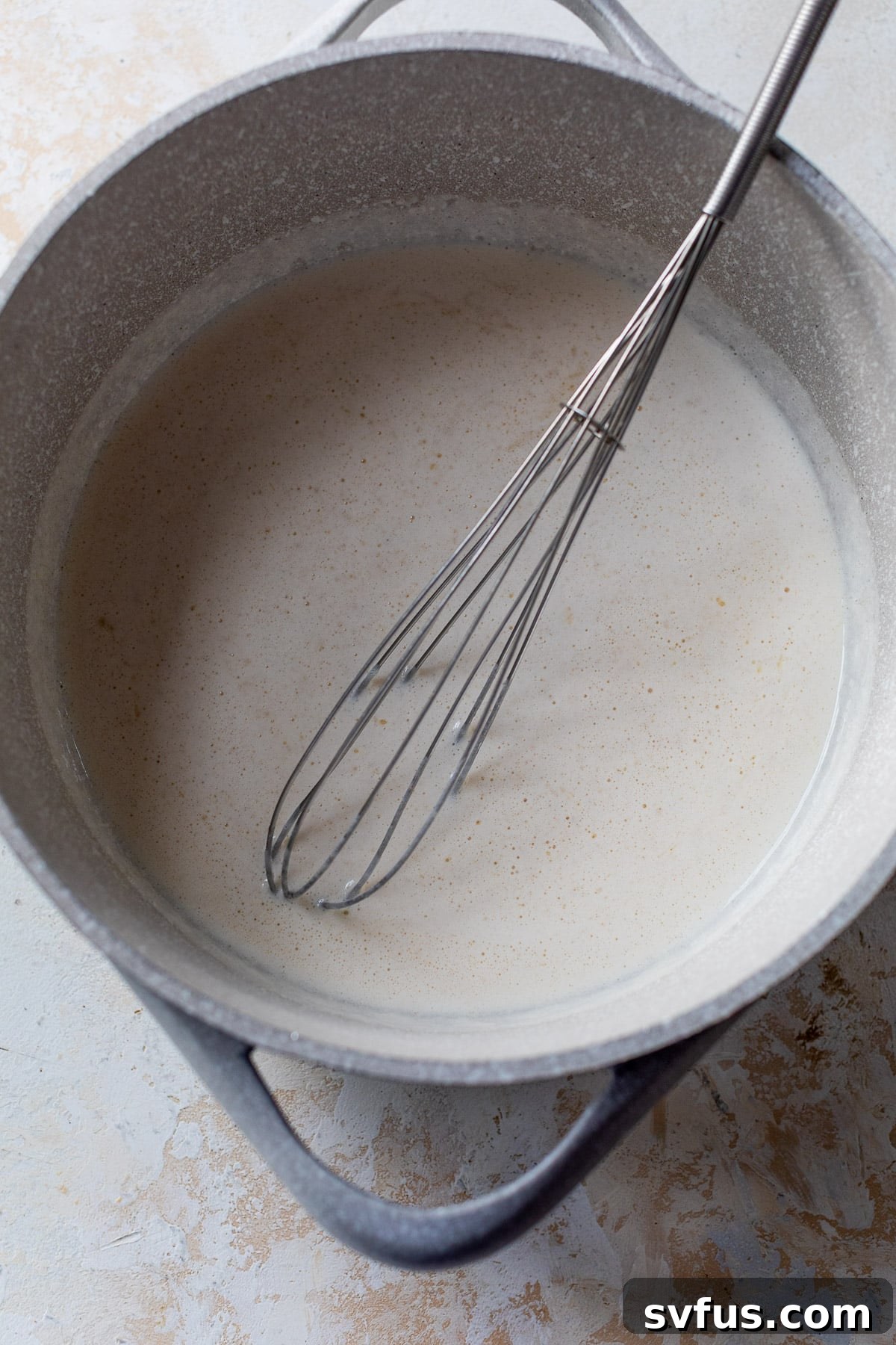 whisking white cream sauce in a large pot, ensuring a smooth consistency before adding cheese.