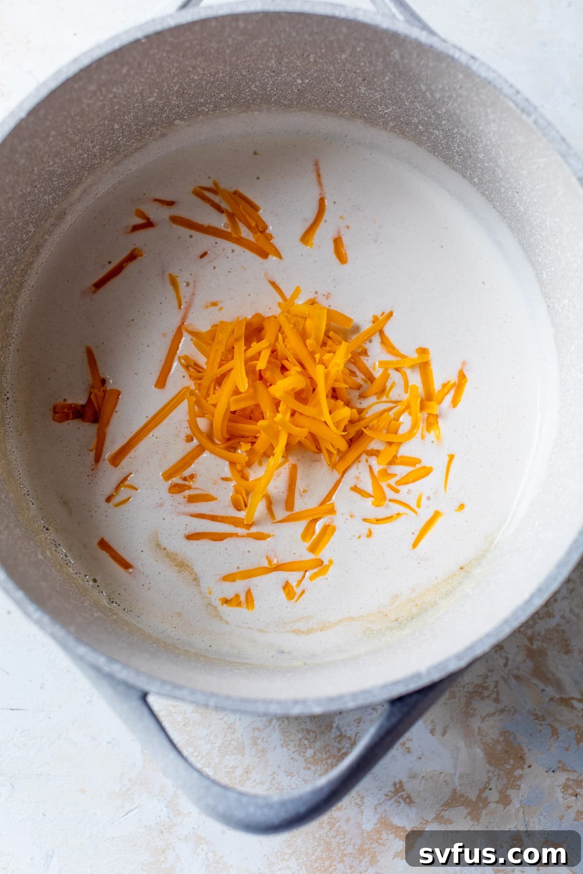 shredded cheddar cheese being slowly added to a pot of creamy white sauce, melting beautifully.