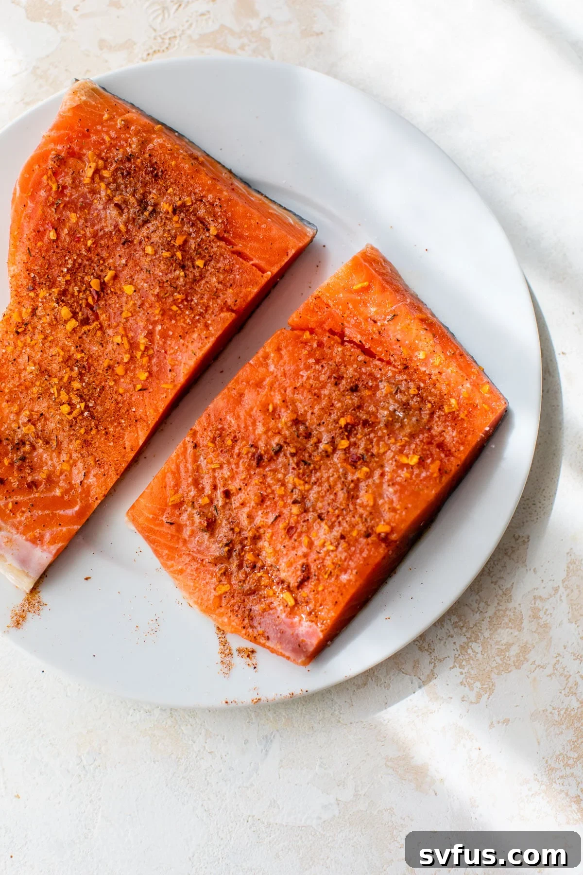 Two raw salmon fillets on a white plate, seasoned generously with lime juice, chipotle seasoning, and salt.