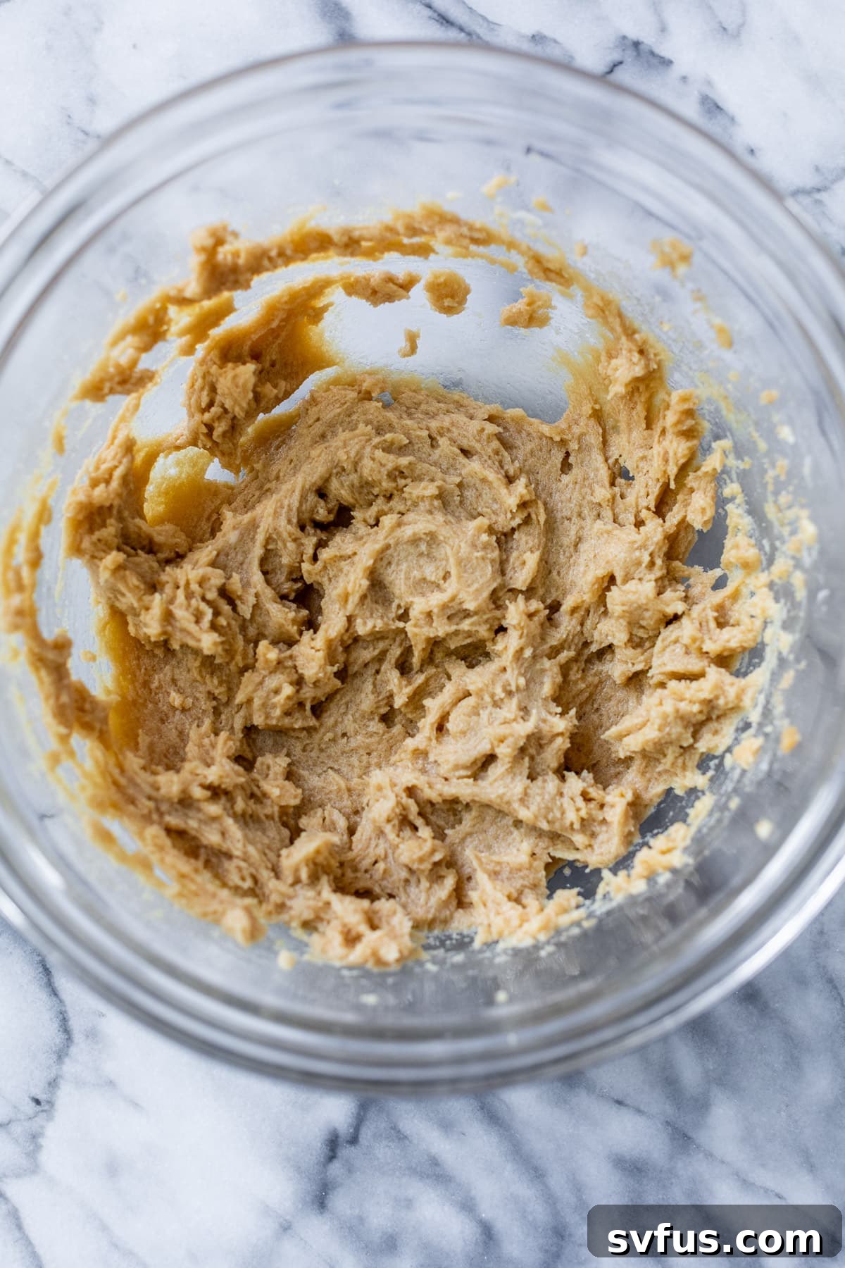 The wet batter for apple oatmeal cookies, a creamy mixture of butter, brown sugar, milk, and vanilla, is ready in a clear glass bowl.