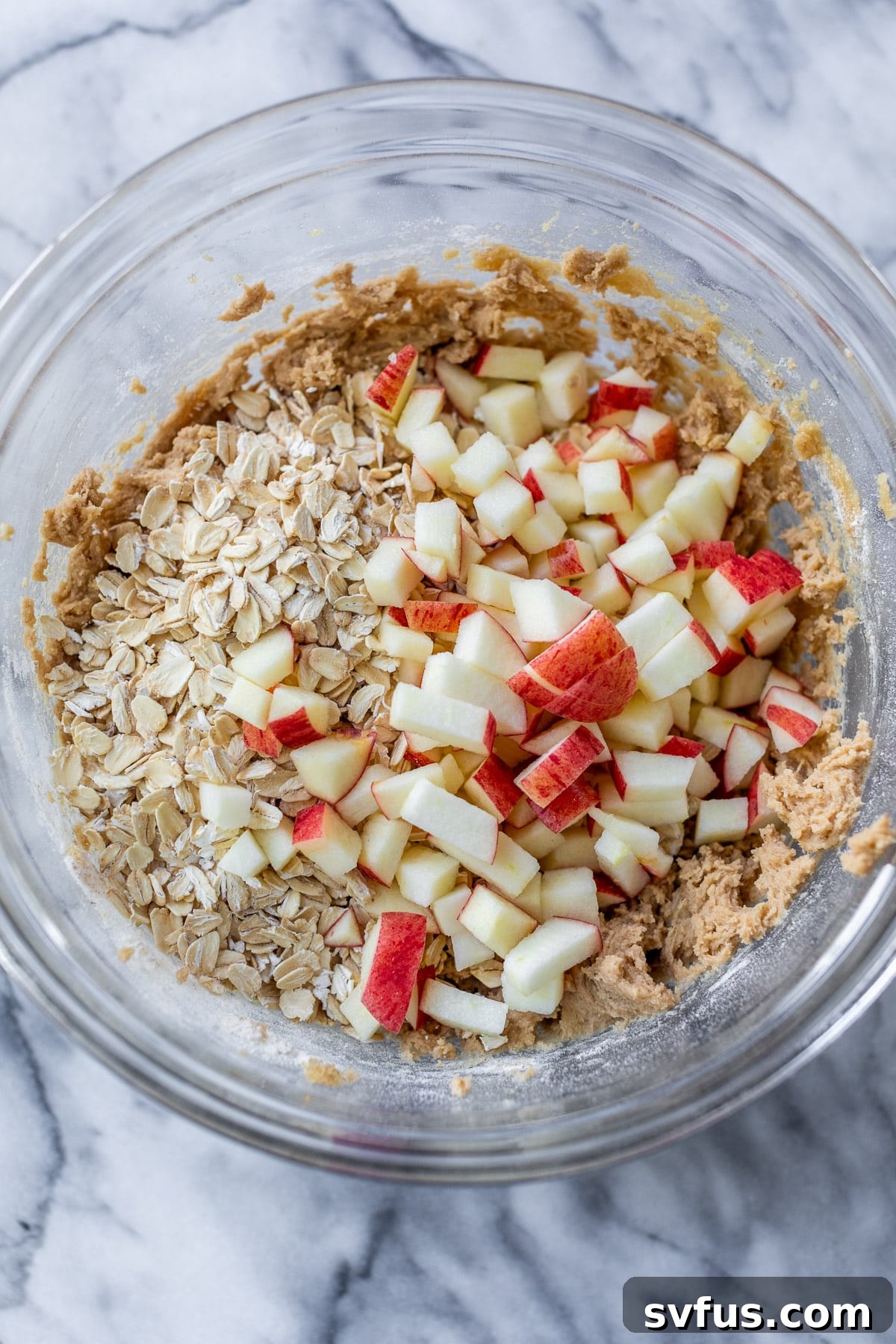 Chopped apple chunks and rolled oats are carefully placed over the top of the mixed apple oatmeal cookie dough in a glass bowl, ready to be folded in.
