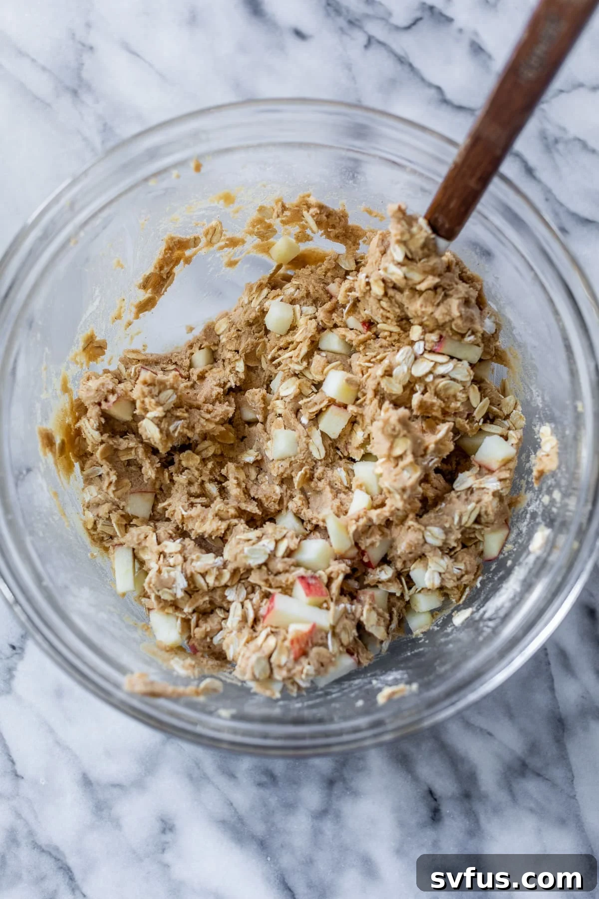 A spatula is used to gently fold oats and apple chunks into the apple oatmeal cookie dough, ensuring even distribution.