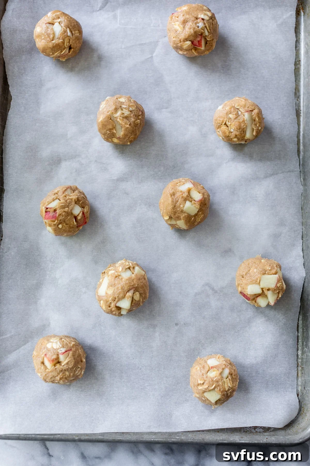 Rolled unbaked apple oatmeal cookie dough balls are neatly arranged on a parchment-lined baking sheet, ready for the oven.