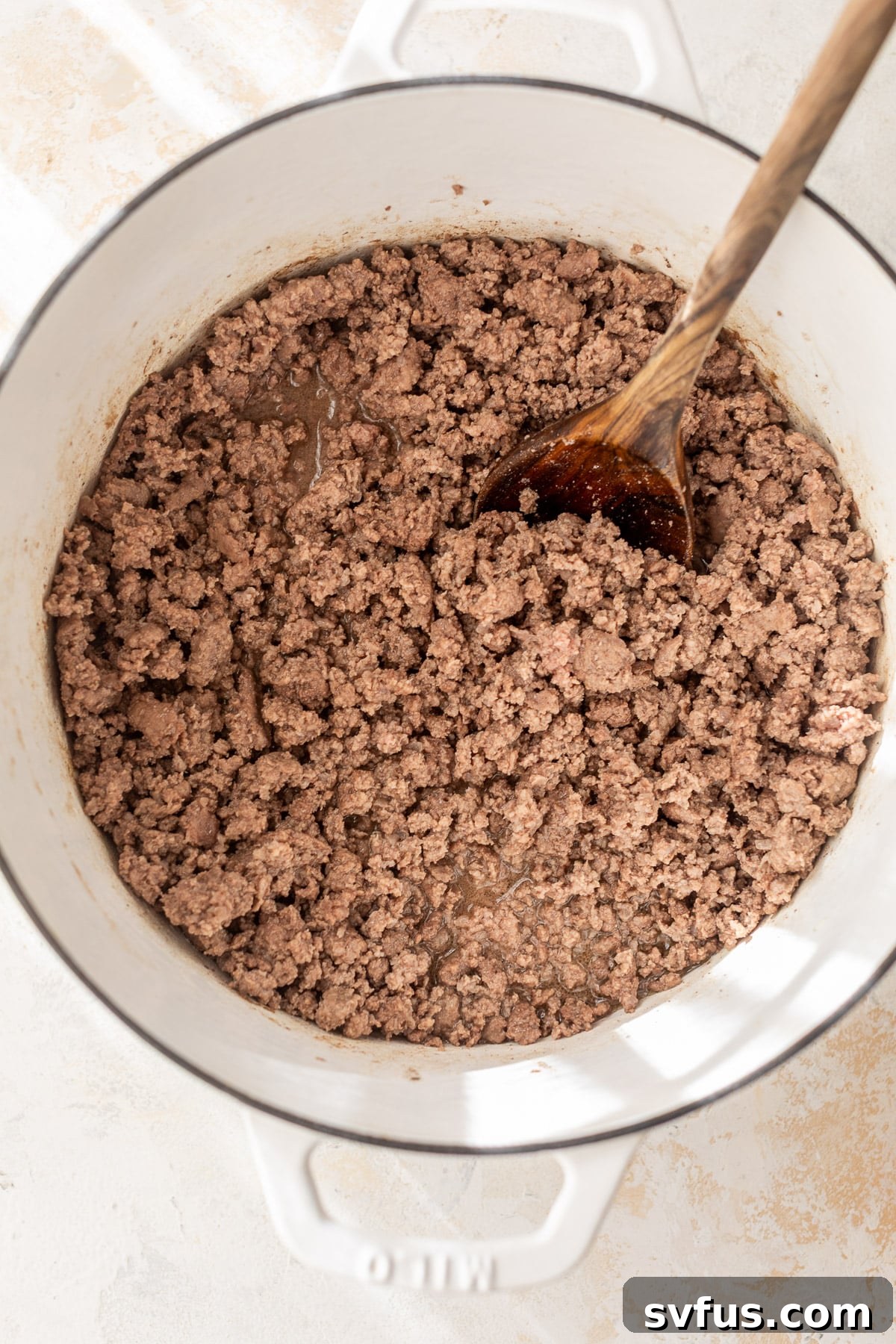 Cooked ground beef in a white Dutch oven, being broken apart with a wooden spoon.