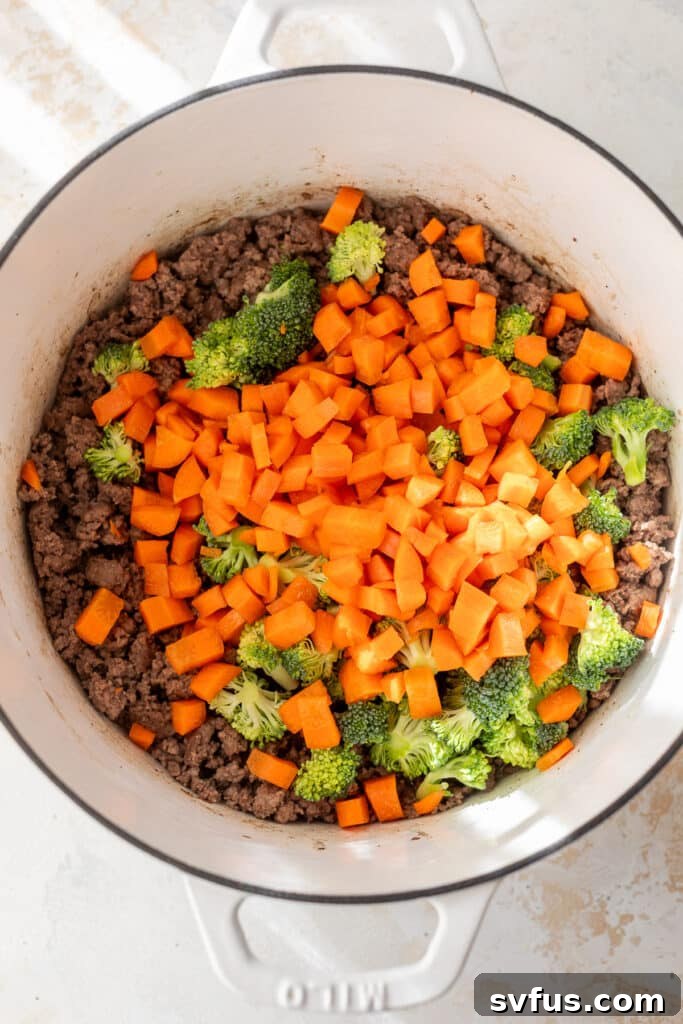 Cooked ground beef with uncooked diced carrots and chopped broccoli in a white Dutch oven.