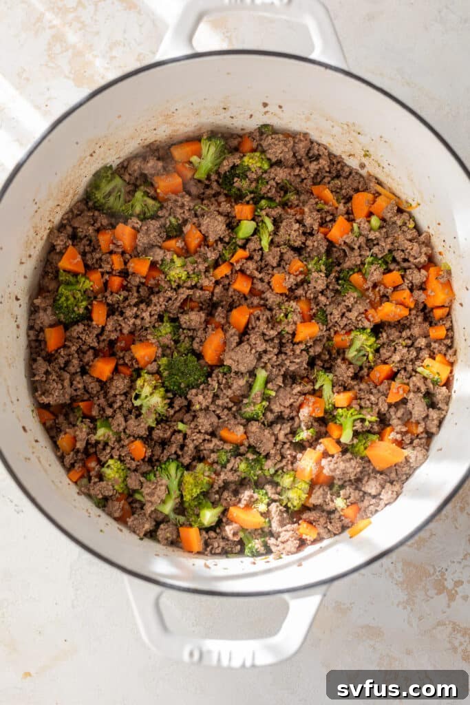 Cooked ground beef, carrots, and broccoli simmering in a white Dutch oven.