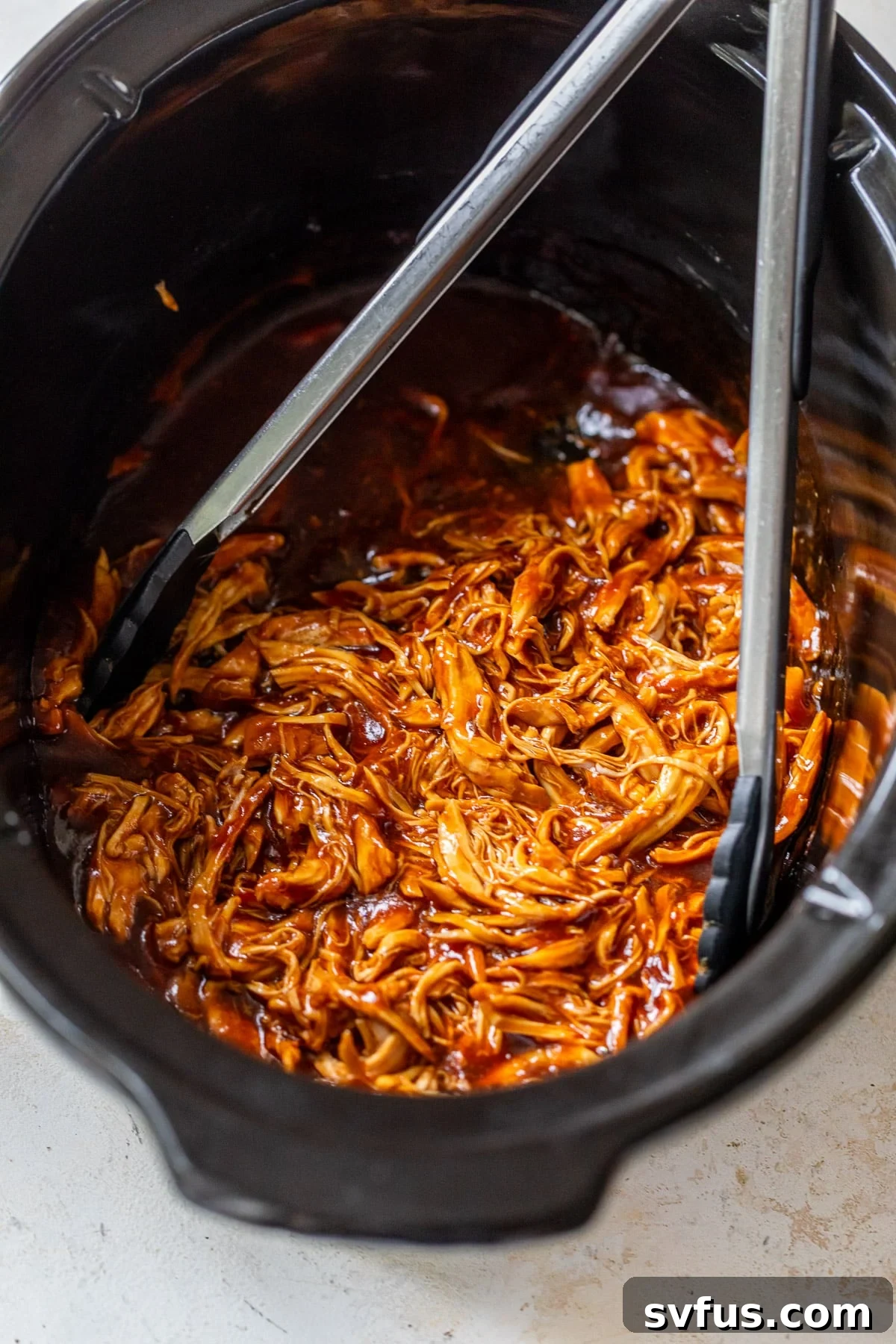 Metal tongs inside a crockpot with shredded BBQ chicken, ready to be served.