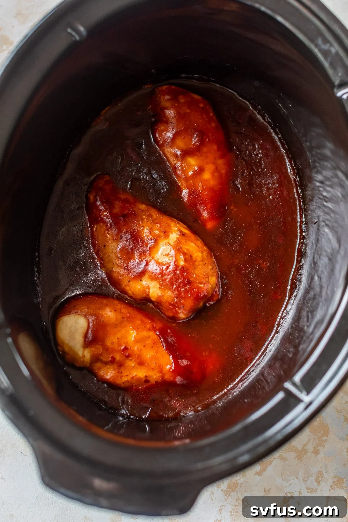 Overhead view of three cooked chicken breasts, glistening with BBQ sauce, after slow cooking in a crockpot.