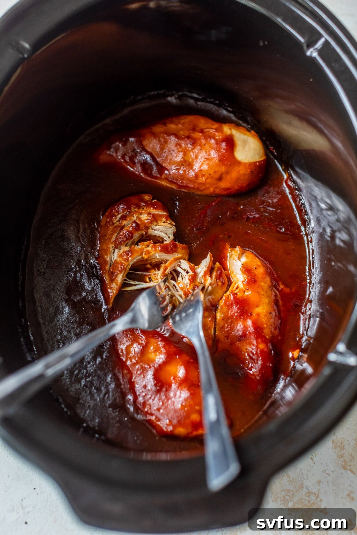 Overhead view of two forks actively shredding tender BBQ chicken breasts directly in a crockpot, surrounded by sauce.