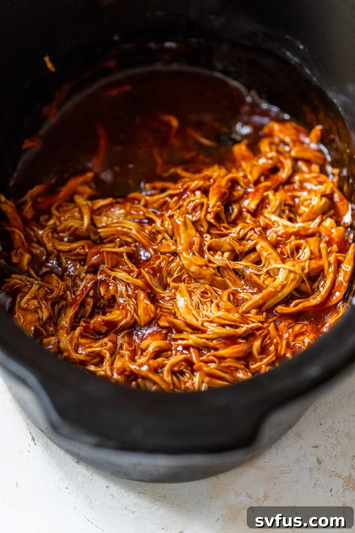 Close up on perfectly shredded BBQ chicken, glistening with sauce, resting in a crockpot.