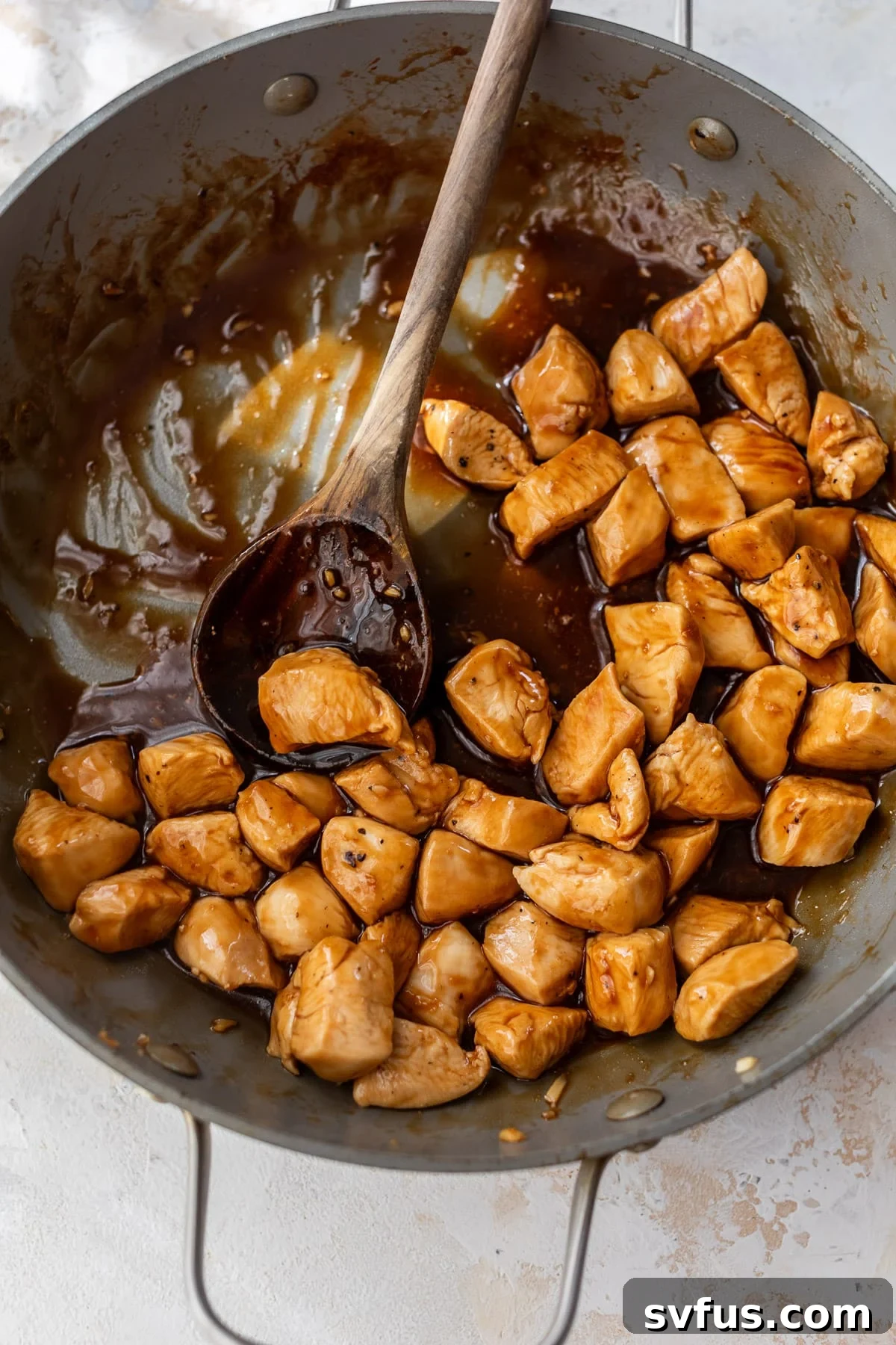 Overhead view of a wooden spoon stirring teriyaki sauce-covered cooked chicken bites in a large skillet.