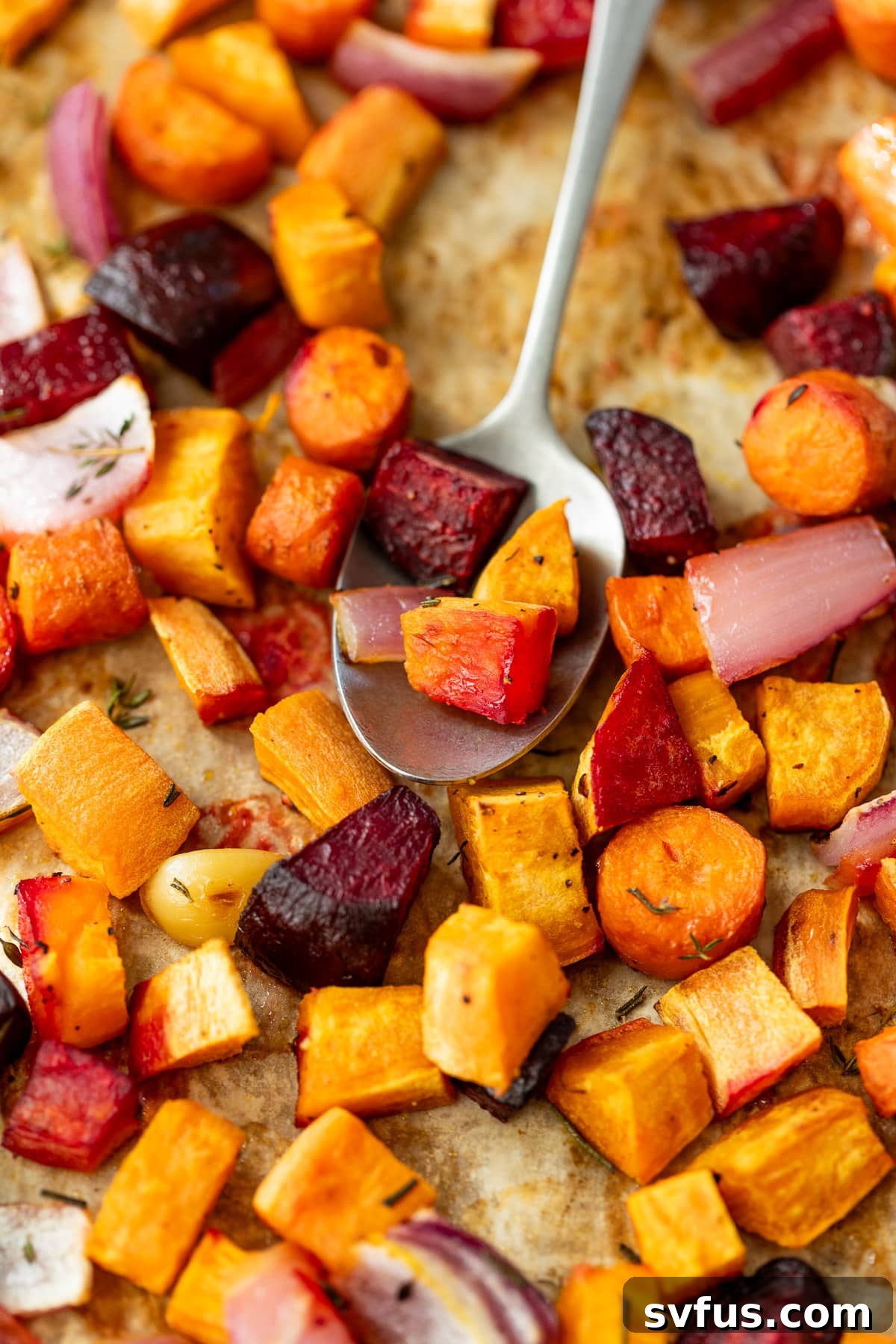 Close-up on a serving spoon resting among a vibrant medley of roasted root vegetables still on the parchment-lined sheet pan.