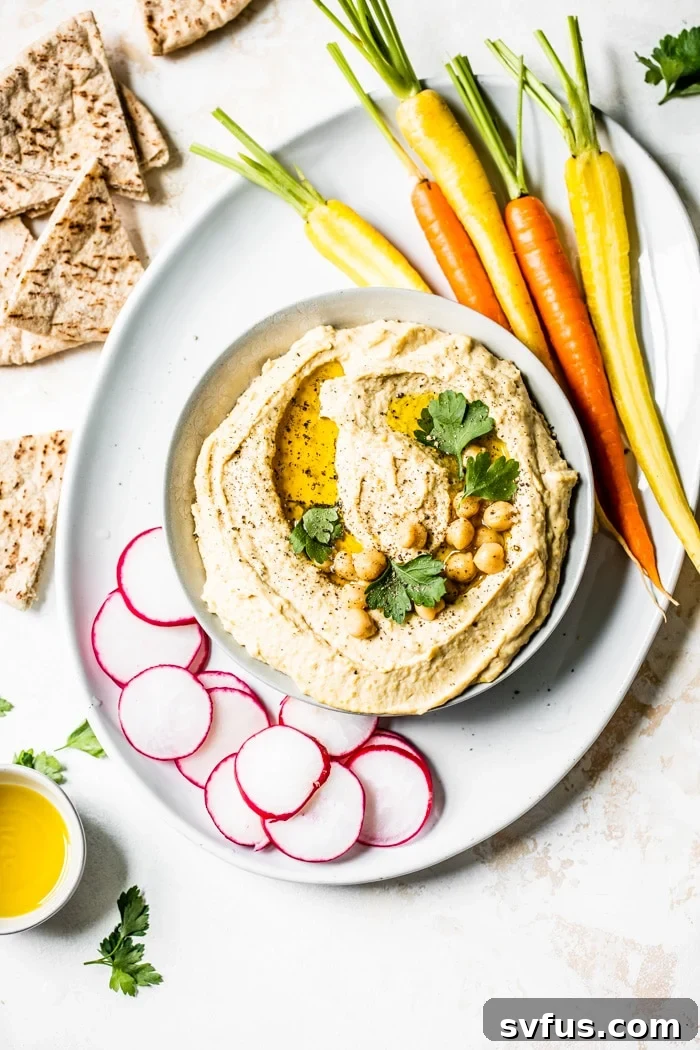 A bowl of creamy homemade hummus topped with whole chickpeas and a drizzle of olive oil, surrounded by fresh vegetables like carrots, cucumber, and bell peppers, along with warm pita bread.