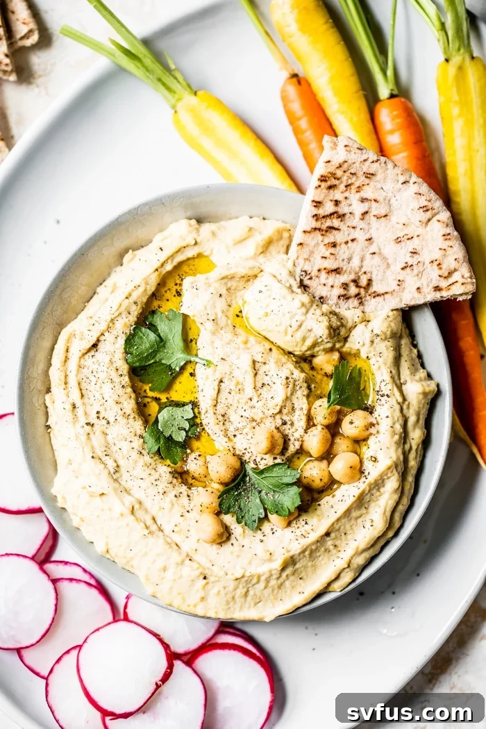 A beautifully presented bowl of creamy hummus, garnished with whole chickpeas, a swirl of olive oil, and fresh parsley, served alongside warm pita bread and a selection of crunchy raw vegetables like carrots and cucumber slices.