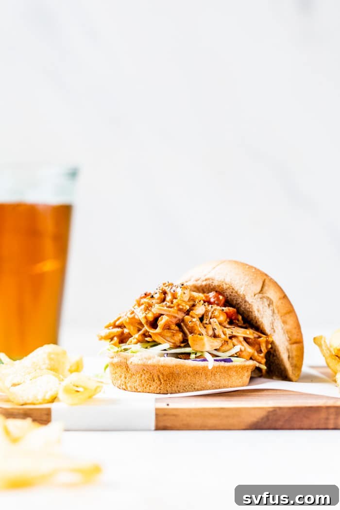 A perfectly assembled hamburger bun filled with savory pulled jackfruit, served alongside crispy potato chips and a refreshing beer, highlighting a delicious vegan meal