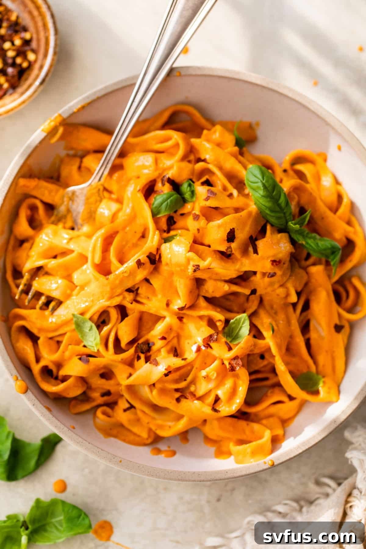 A beautifully plated bowl of long vegan roasted red pepper pasta with a fork twirled around it, garnished with fresh herbs.