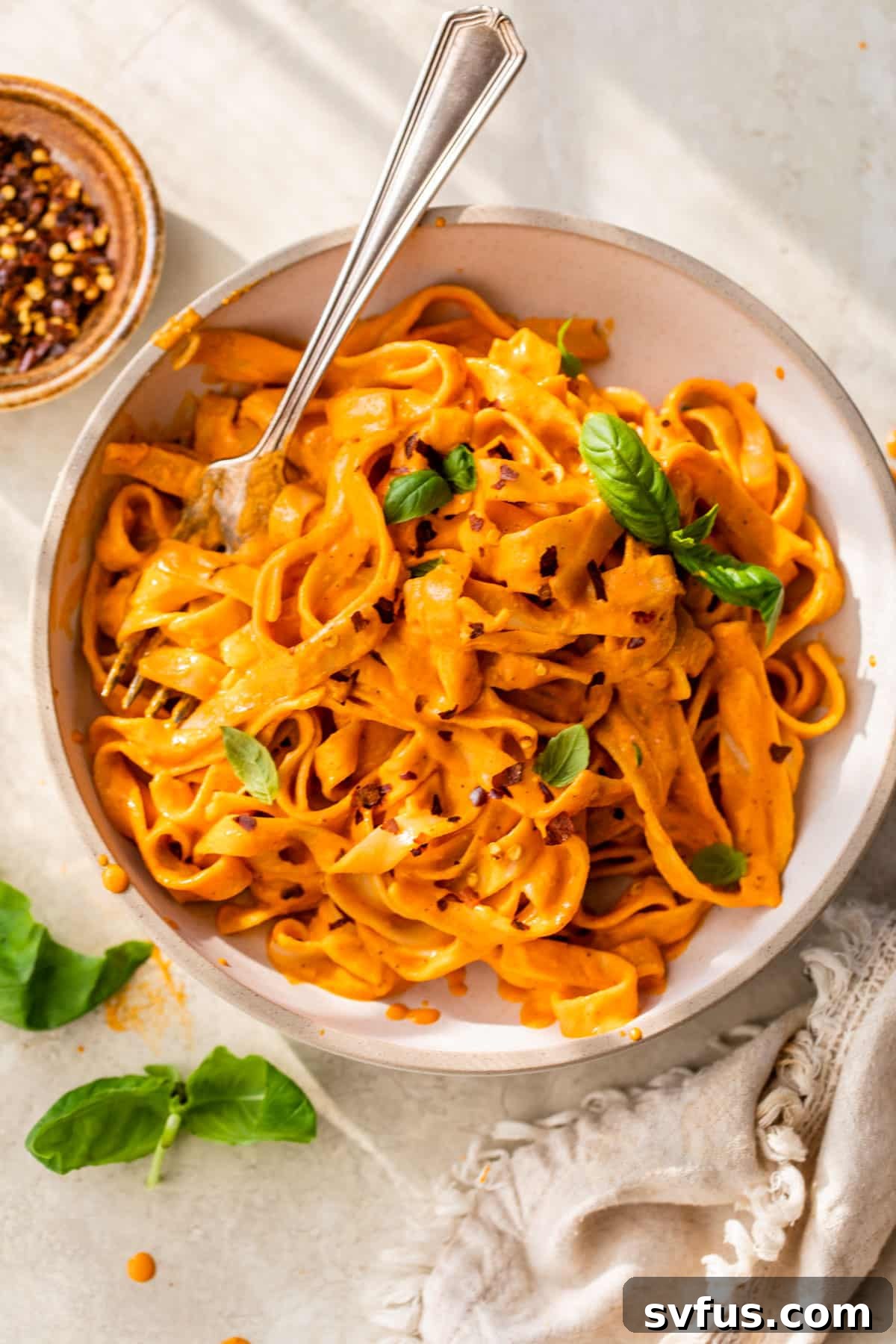 A vibrant bowl of vegan roasted red pepper pasta, generously topped with fresh green basil leaves, creating a beautiful contrast of colors.