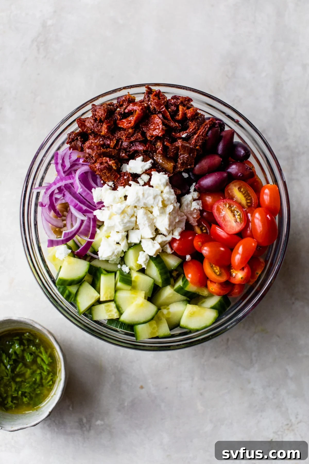 A vibrant bowl filled with cooled pasta, cucumber, tomatoes, red onion, sun-dried tomatoes, and feta cheese