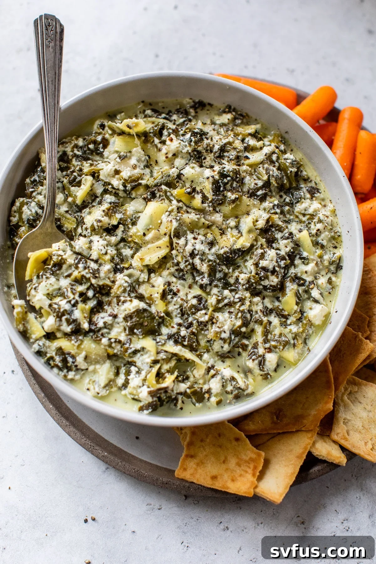 A beautifully garnished bowl of Crockpot Spinach Artichoke Dip, surrounded by fresh pita chips and vibrant baby carrots, ready for serving.