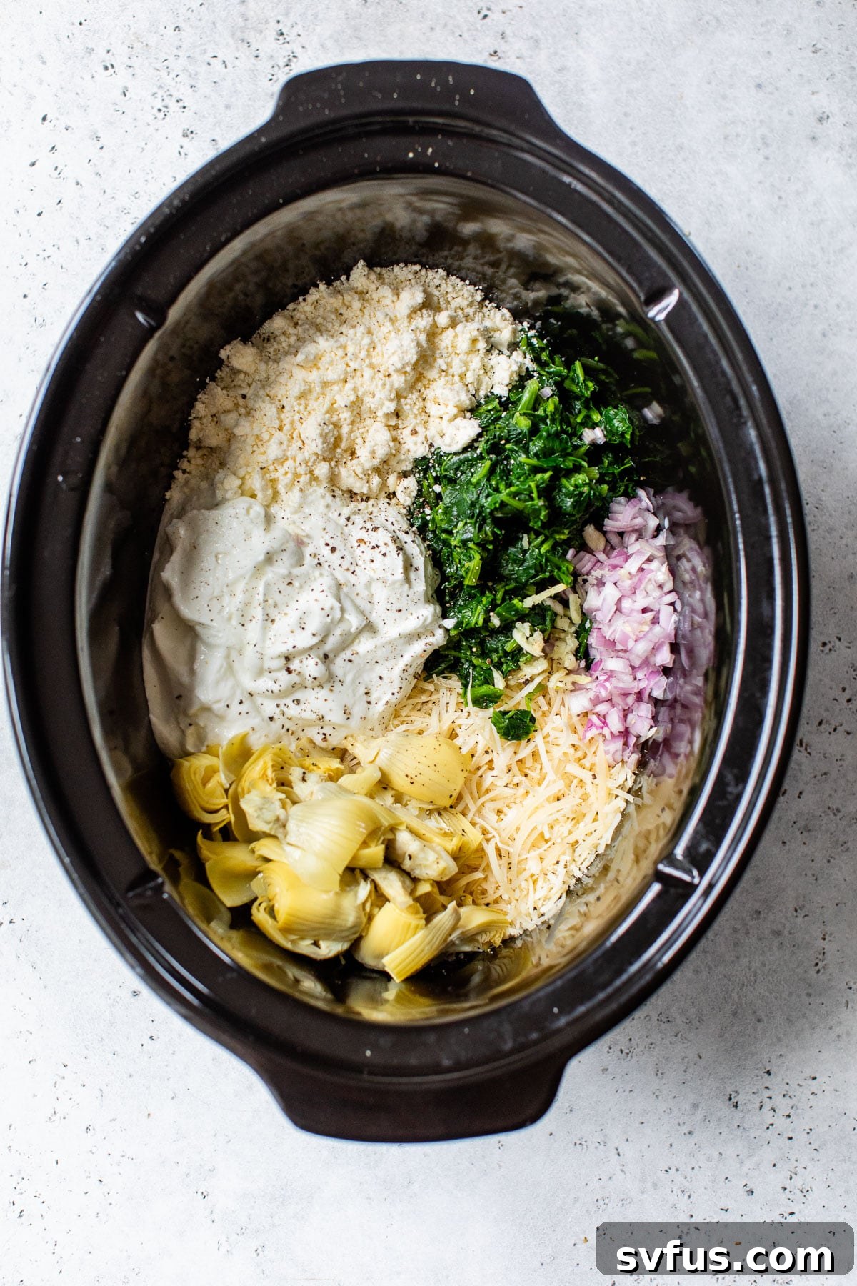 A slow cooker bowl filled with the raw ingredients for spinach artichoke dip, including feta, spinach, shallots, parmesan, artichokes, and Greek yogurt, before cooking.