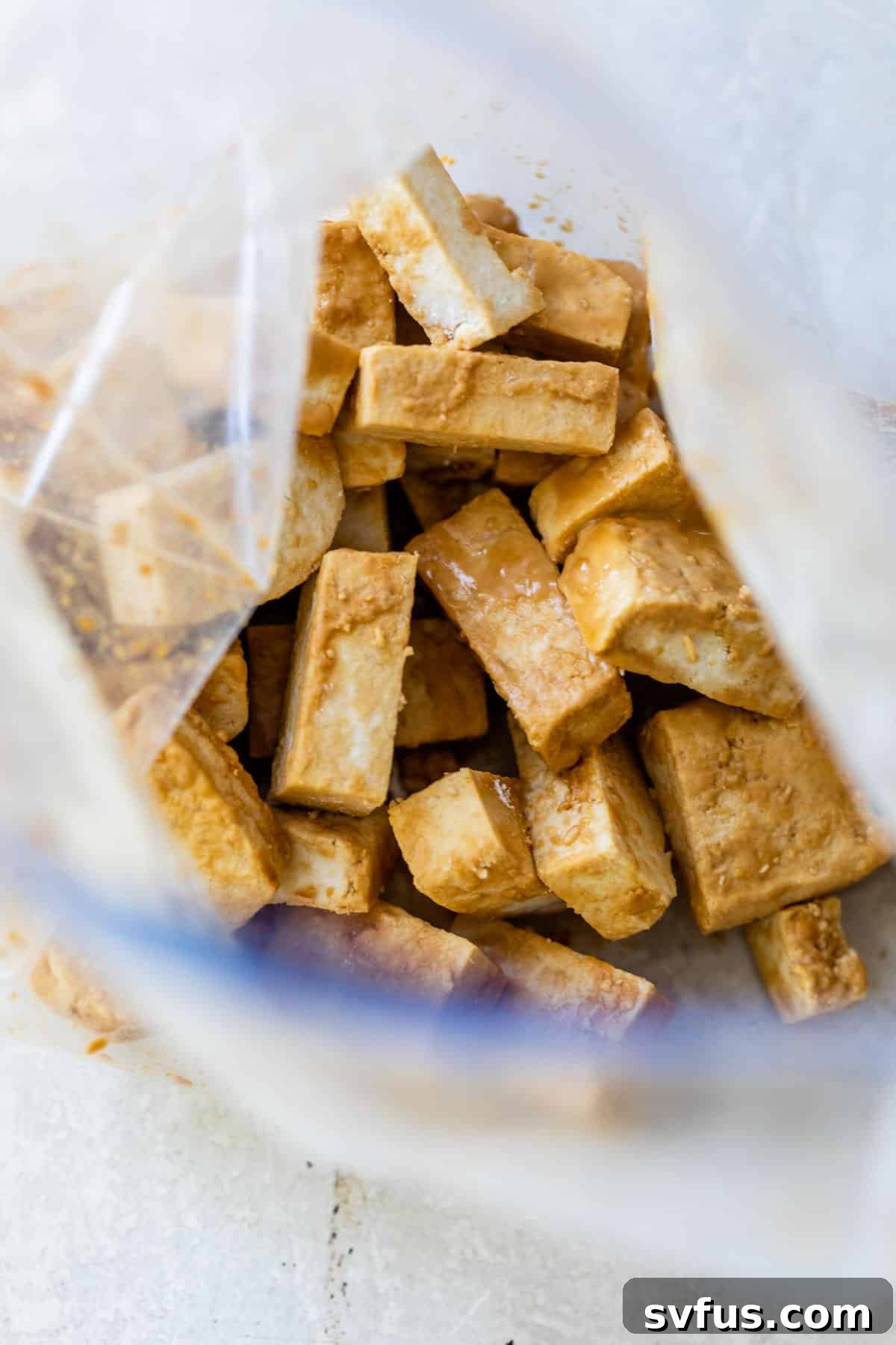 Cubed tofu pieces coated in soy sauce and cornstarch, marinating in a clear plastic bag, ready for pan-frying to achieve optimal crispiness.