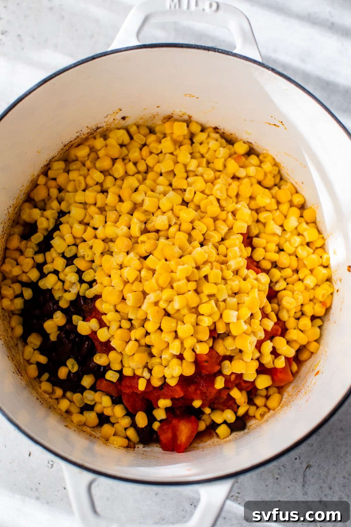 Corn, black beans, diced tomatoes, and vegetable broth added to the sautéed aromatics in a Dutch oven, ready to be stirred.