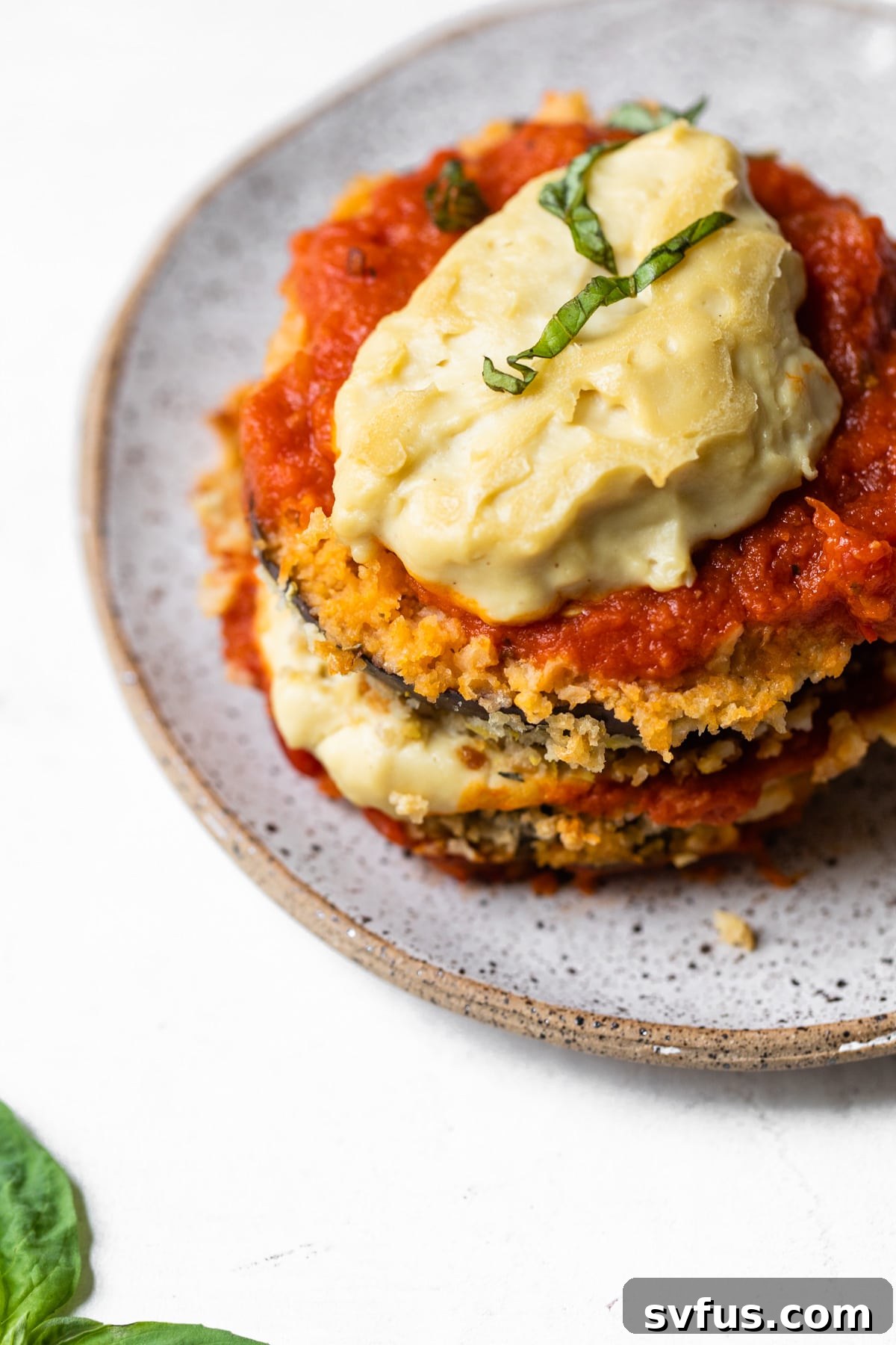 A close-up of a single serving of Vegan Eggplant Parmesan on a white plate, garnished with fresh basil.