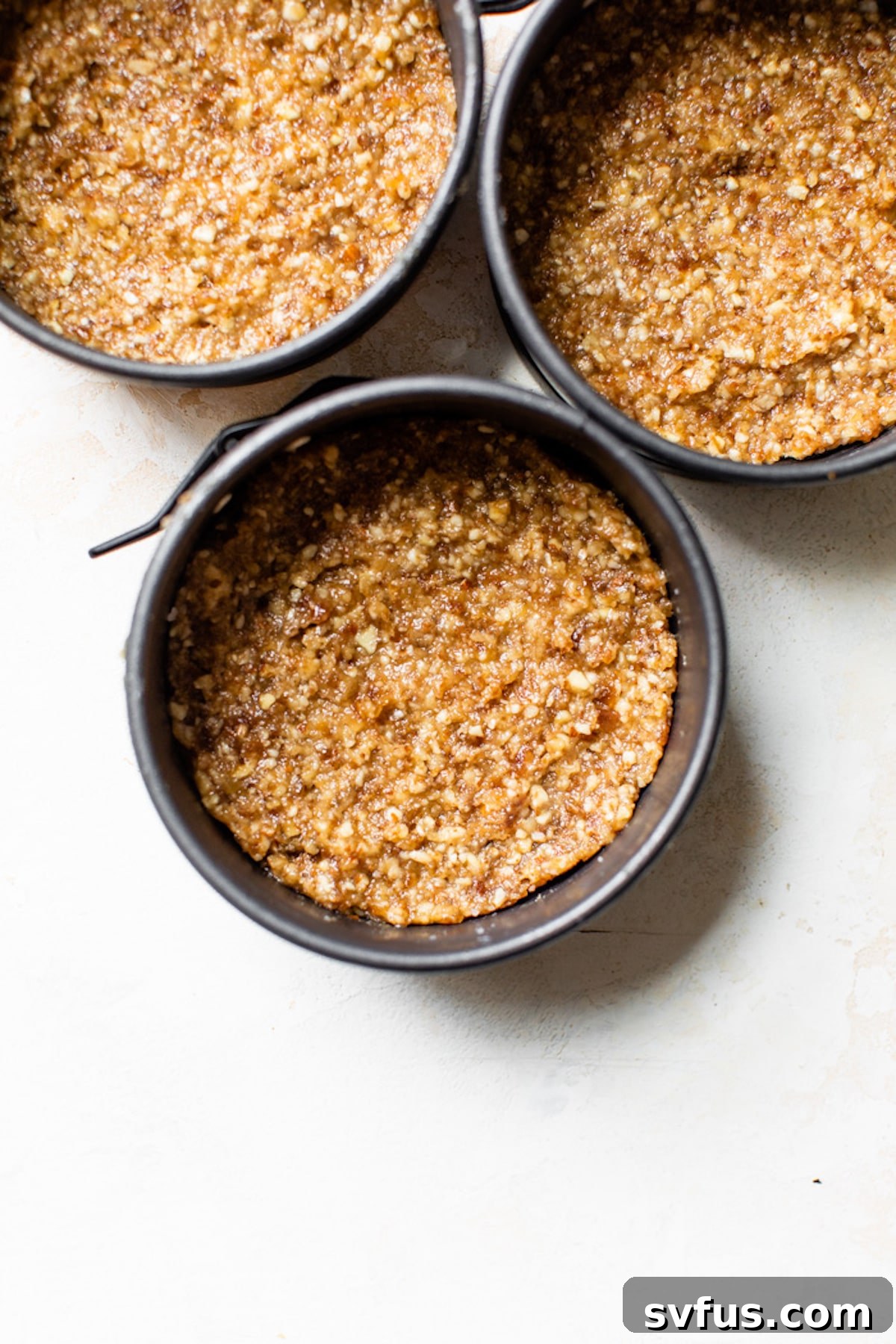Four small 4-inch springform pans, each filled with a pressed nut-based crust, ready for the cheesecake filling.